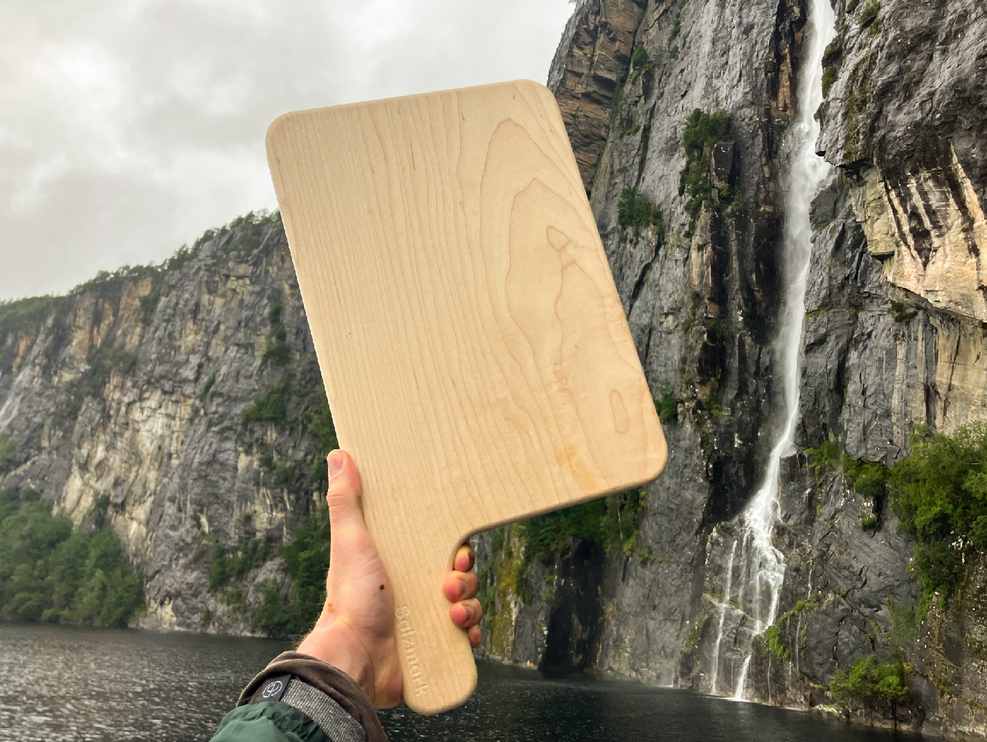 Hand holding a solid wooden cutting board with a waterfall and mountainous landscape in the background