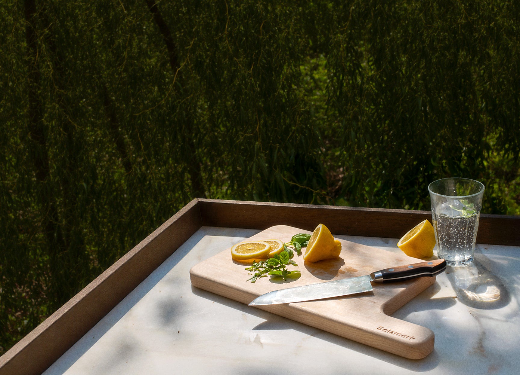 Wooden cutting board with lemons, a knife, and a glass of water on a marble surface with a blurred green outdoor background.
